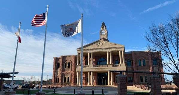 City hall building in Roanoke, TX. Large brick rectangular building with tan stone columns in front and clock tower on top.