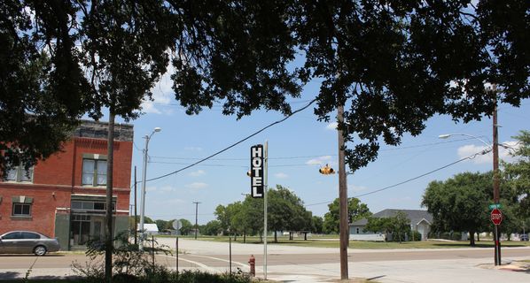 An intersection in Coppell with tree overhead and black sign that says "Hotel" vertically. Brick building across street on left.