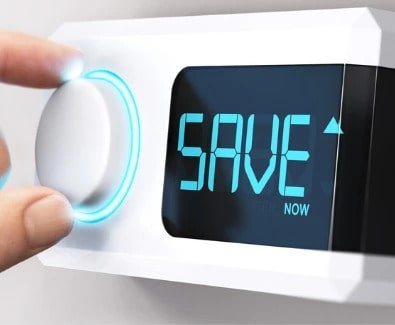 Person's hand adjusting a knob on a white thermostat on a wall. Display screen says "SAVE"