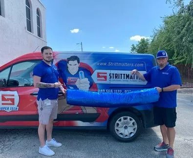 Two technicians holding something long an blue between them, standing in front of a service van in a parking lot.