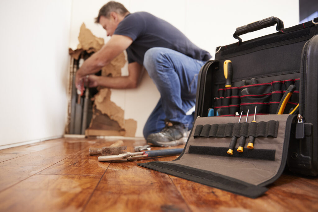 Plumber repairing a leaky pipe through a hole in a white wall. Open tool bag in foreground, on wood floor.